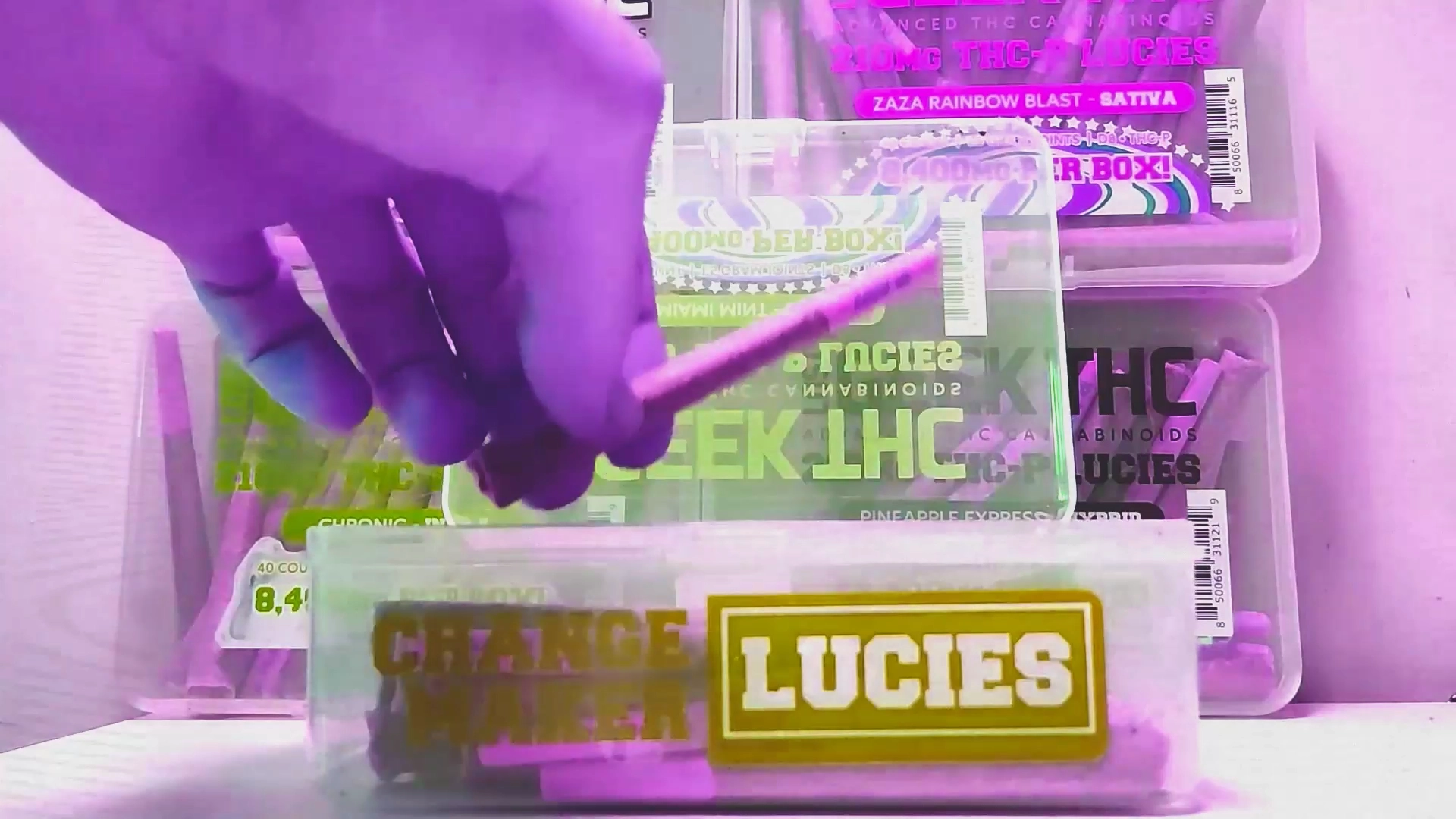 A purple-tinted scene showing a hand holding a cannabis pre-roll above a translucent plastic container labeled ‘LUCIES,’ with blurred Geek THC Lucies packaging visible behind it.